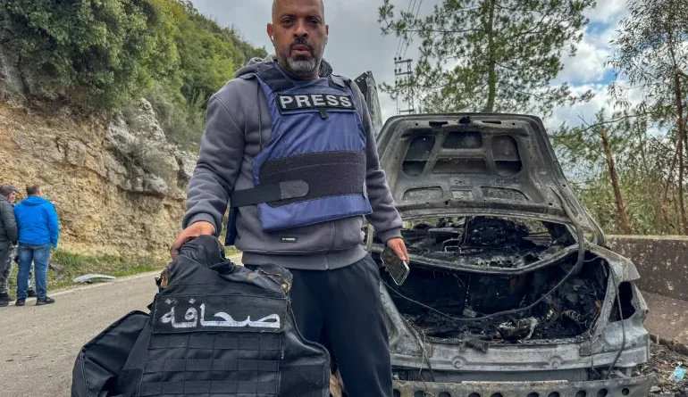 يحمل الصحافي جمال الغرابي من قناة الميادين سترة صحافية إلى جانب السيارة التي دُمّرت جراء غارة إسرائيلية استهدفتها وأودت بحياة زملائه [علي هنكير/رويترز].